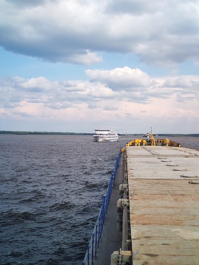 A wide-angle, eye-level shot captures a serene river scene under a partly cloudy sky. In the foreground, the weathered deck of a barge extends from the right edge of the frame, its surface showing signs of age and use with a light brown, textured appearance and visible seams and metal fittings. A blue railing runs along the left edge of the barge, separating it from the dark, rippling water of the river. In the middle distance, a white passenger ship, with multiple decks and windows, sails away from the camera towards the left. The ship appears to be navigating the broad river, with a line of green trees forming a distant treeline on the horizon. The sky is a mix of pale blue and white, with scattered cumulus clouds tinged with pink and grey, suggesting it might be late afternoon or early evening. The overall impression is one of peaceful transit on a wide waterway. The location is the city of Krokhino, Russia, a place historically significant for its shipbuilding and river trade.