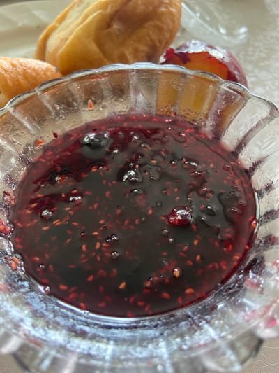 A close-up shot shows a bowl of dark red jam, possibly raspberry or blackberry, with visible seeds and some whole berries. The jam is in a clear, fluted glass bowl. In the background, partially visible, are pieces of baked goods, likely bread or pastries, and a slice of plum. The setting appears to be a table with a textured tablecloth, possibly in a home or cafe. The image does not provide any specific information to determine its location in Askarovo, Russia.