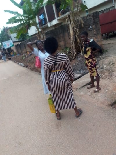 This image captures an outdoor scene in Orlu, Nigeria, featuring three women on what appears to be a paved road or pathway. The setting suggests a semi-urban or residential area, with buildings and vegetation in the background.
In the foreground, a woman faces away from the camera, wearing a long dress with distinct vertical black and off-white stripes, cinched at the waist with a thin yellow tie or belt. She has dark, curly hair and carries a light green or yellow reusable bag on her left arm. She is wearing dark flat sandals.
To her left and slightly behind, another woman is partially visible, facing towards the right side of the frame. She wears a light-colored, possibly white or light blue, striped top and has her hands on her head, suggesting a moment of thought, exasperation, or adjusting her hair. She carries a red plastic bag on her arm and wears glasses.
On the right side of the frame, a third woman stands, looking towards the other two. She is dressed in a vibrant yellow and dark-patterned sleeveless dress, with a dark garment (possibly a shawl or light jacket) draped over her shoulders. She wears light-colored flat sandals.
The background shows a mix of natural and built environments. Lush green foliage, including large leaves resembling banana or plantain trees, is prominent. Behind the women, there's a low, rough-textured concrete wall. Further back, multi-story buildings with balconies are visible, with some laundry hanging. To the far left, a blurred sign with colorful circular graphics and some text, likely a public information notice, can be seen. The road surface is granular and light-colored. The soft, even lighting suggests either an overcast day or the late afternoon/early evening. No specific text is clearly legible.
