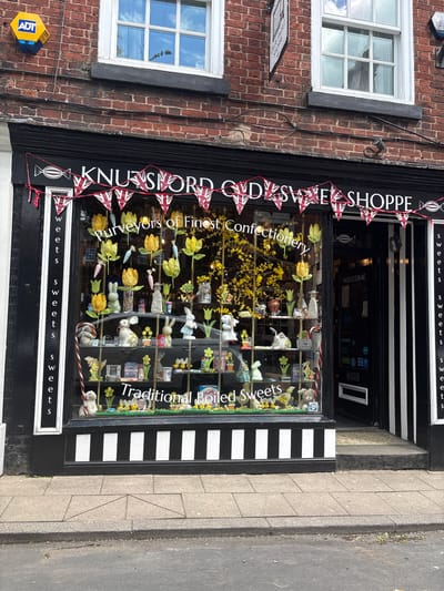 A street-level view presents "Knutsford Olde Sweet Shoppe" in Knutsford, United Kingdom, noted as "Purveyors of Finest Confectionery." The shop facade is black with white lettering. A garland of Union Jack flags stretches across the top of the shopfront. The window display contains an assortment of sweets and floral decorations, including yellow tulips and bunny figurines. Signage within the window reads "Traditional Boiled Sweets." The building's architecture includes brickwork and white-framed windows above the shopfront. An ADT security box is visible on the left side of the building. A portion of the pavement is visible to the right of the shop.