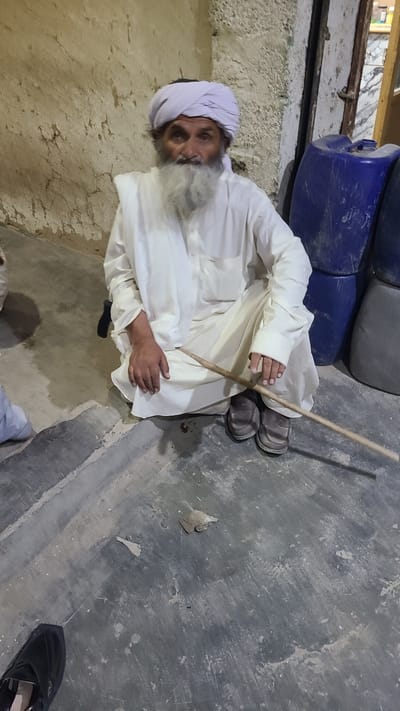 A man with a grey beard and a white turban sits cross-legged on a concrete floor in what appears to be an indoor space. He wears a white outfit and grey shoes. The man holds a wooden stick in his hands, oriented vertically. Behind him are several blue and grey plastic containers. The wall to his left is rough, possibly concrete. The floor also shows signs of wear and discoloration. A black shoe is visible in the lower right corner of the image. The location is Ziarat, Pakistan.