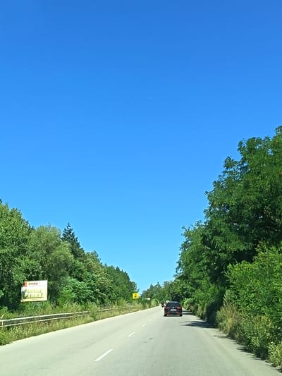 The image depicts a two-lane asphalt road in a rural setting, likely within or near Ruska Bela, Bulgaria. The road curves gently to the left as it recedes into the distance. On the right side of the road, a dense line of lush green trees forms a natural barrier. On the left side of the road, there is a roadside guardrail and further back, more dense vegetation and trees. A billboard with what appears to be text and an image is visible on the left, next to the guardrail. Further down the road, on the right, a yellow triangular road sign with a black symbol is present. A dark-colored SUV is driving away from the camera on the right lane. The sky is a clear, vibrant blue with no clouds. The scene suggests a sunny day with good visibility.