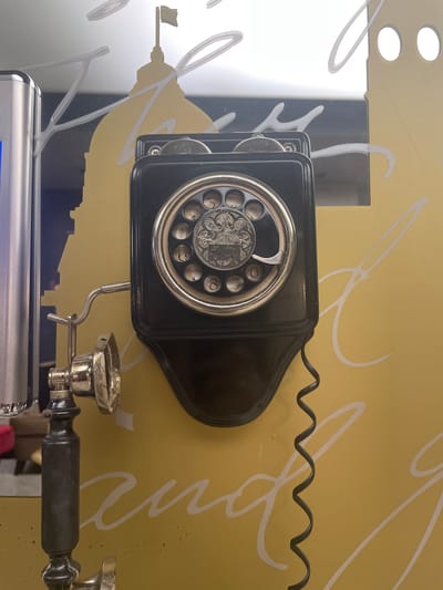 An antique black wall-mounted rotary dial telephone is the primary subject of this image, positioned against a yellow wall decorated with white cursive lettering and the silhouette of a domed building topped with a flag. The telephone's receiver is detached and hangs from a hook, connected to the base by a coiled black cord. The dial features numbers and ornate detailing. To the left of the telephone, a portion of a silver metallic object, possibly a modern appliance or fixture, is visible. The overall scene appears to be an interior, likely a themed establishment, and the background elements suggest a potential connection to a significant landmark in İzmir, Turkey, though the specific location is not explicitly identifiable from the image alone.