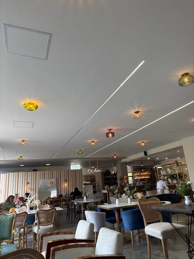 The image depicts the interior of a well-lit establishment, possibly a cafe or restaurant, in Rionegro, Colombia. The scene is captured from a low angle, looking upwards towards a white ceiling adorned with multiple stylish light fixtures. These fixtures are glass sconces that emit warm, patterned light, casting intricate shadows on the ceiling. Two long, thin LED light strips run parallel across the ceiling, adding to the modern aesthetic.

The background reveals a bustling space with various seating arrangements. There are tables and chairs, some with upholstered seats in blue and cream, and others made of woven rattan. Shelving units display decorative items, including vases and what appear to be ceramic plates. In the distance, several people can be seen interacting within the space, with one individual in the background appearing to be a staff member.

The overall atmosphere seems relaxed and inviting, suggesting it could be daytime or early evening due to the bright interior lighting. The decor incorporates a mix of modern and possibly vintage elements, with the patterned wallpaper in the background adding a touch of vibrancy. A neon sign with the word "Kitchen" is visible in white text against the wall, clearly indicating a food service area. The image conveys a sense of a social gathering or dining experience within a thoughtfully designed environment.