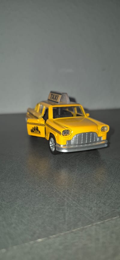A yellow toy taxi, resembling a vintage Checker Cab, is positioned on a dark, textured surface. The taxi has its driver-side door ajar, revealing a black and white checkered interior trim with a graphic of city buildings. A "TAXI" sign is mounted on the roof. The background is a plain, light gray wall. There are no people or animals present. The image provides no information about the city of Baden-Baden, Germany.