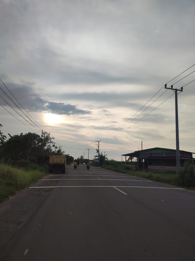 The image depicts a straight asphalt road in Keureusek, Indonesia, stretching into the distance under a cloudy sky with the sun obscured. A yellow dump truck is parked on the left side of the road, and two motorcycles are visible further down the road. Power lines run parallel to the road above. Vegetation lines the left side of the road, and a building sits on the right. Yellow and white markings are present on the road surface.