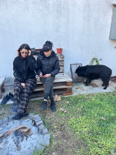 A photo taken in Florida, Argentina depicts two people, a black dog, and a fire pit in an outdoor setting. The two individuals are seated on a makeshift wooden pallet bench. One person is wearing a black jacket and dark pants, while the other is wearing a dark jacket and plaid pants. A black dog stands near the bench on a concrete area adjacent to a grassy patch. A fire pit constructed with gray stones contains wood and is emitting flames. The area is surrounded by a white wall and some greenery.