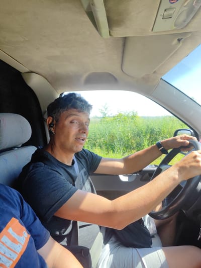 A man with short, dark hair and a light complexion is driving a vehicle, his left hand on the steering wheel. He is wearing a dark t-shirt and a grey seatbelt. A black wireless earbud is in his left ear. In the foreground to the left, another person is visible from the shoulders down, wearing a blue t-shirt with an orange graphic. The interior of the vehicle is visible, including the ceiling with a sun visor and a light fixture. The exterior environment, seen through the windshield, is a landscape of green vegetation under a clear blue sky. The image appears to be taken from inside the vehicle, looking towards the driver.