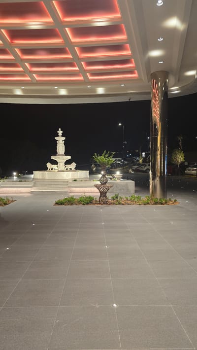 The image depicts an outdoor nighttime scene at what appears to be the entrance to a modern establishment, possibly a resort or entertainment venue, in Agios Epiktitos, Northern Cyprus. The foreground is dominated by a large expanse of grey tiled flooring, with several small, circular lights embedded in it. In the mid-ground, a multi-tiered white marble fountain is the central focus. The fountain features sculpted figures and lion motifs, and it is illuminated from below. To the right of the fountain is a decorative metal structure with a potted plant. Behind the fountain, a parking area with several cars is visible, illuminated by streetlights. The scene is framed by an overhead structure with a ceiling featuring a grid of recessed lighting panels emitting a warm red glow. A large, polished metal column stands prominently on the right side of the image, reflecting the ambient light and the red ceiling illumination. The background is a dark night sky, with some distant lights suggesting the presence of buildings or a town. There are no people visible in the frame. The overall impression is one of an elegantly designed and well-lit entrance, designed to impress visitors.