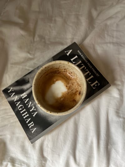 An overhead shot captures a cup of coffee with foam resting on a book titled "A Little Life" by Hanya Yanagihara. The book is placed on a white, rumpled bedsheet. The scene is devoid of any individuals or specific city markers, making it impossible to ascertain its location in Aarhus, Denmark, or any other city.