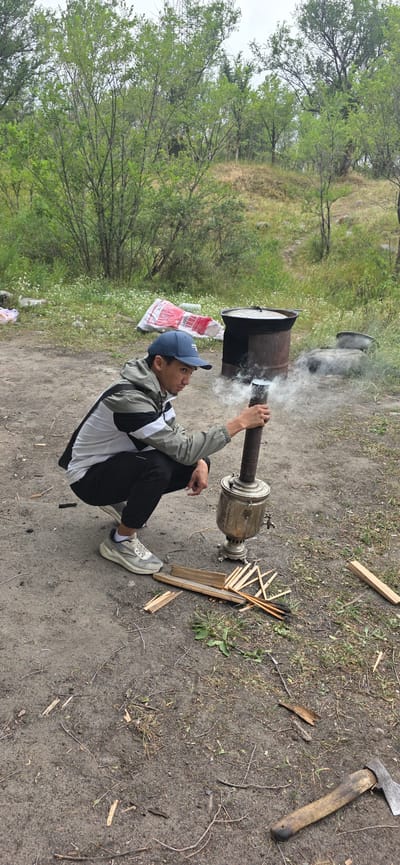 A young man, wearing a blue baseball cap, a gray and black jacket, and black pants, is sitting on the ground in an outdoor setting. He is holding a long metal pipe connected to a samovar. Smoke is emanating from a large metal container positioned behind him, suggesting the samovar is being heated. He is looking towards the container.
The scene appears to be a rustic, natural environment, likely in Кашка-Суу, Kyrgyzstan, with trees and vegetation visible in the background. The ground is a mix of dirt and sparse grass, with some scattered wood pieces. To the right of the samovar, several pieces of firewood are piled up, along with an axe. In the background, a large black pot is also visible, with smoke rising from it. The lighting suggests it might be daytime, possibly overcast. The overall impression is of a person engaged in preparing a meal or a hot drink outdoors.
