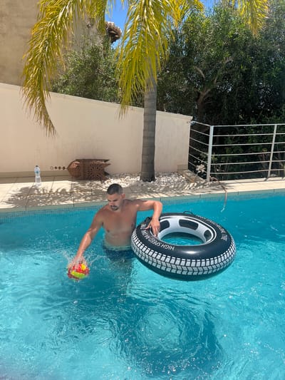 A medium shot captures a man in a swimming pool, potentially in a residential area of El Poble Nou de Benitatxell/Benitachell, Spain. He is partially submerged, wearing what appears to be blue swimming trunks, and is holding a brightly colored ball in his right hand, creating a splash. The man is also using his left arm to hold onto a black and white inflatable tire.
To the left, the swimming pool is bordered by a cream-colored wall and tiled edge. Beside the wall are decorations, a water bottle, and white pebbles. Above the wall, foliage and trees are visible. A palm tree with yellow leaves is positioned to the left, partially obscuring the scene. Sunlight illuminates the scene.