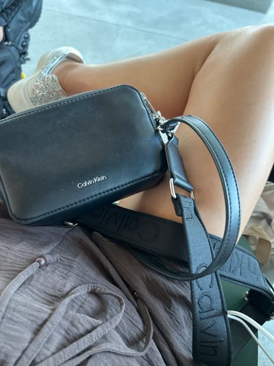 The image shows a close-up of a person's legs, wearing a sparkly silver slip-on sneaker and sitting down, with a black Calvin Klein crossbody bag resting on their lap. The bag is made of faux leather and has a silver zipper. The strap of the bag is black with repeated Calvin Klein logos. The person is wearing a casual, loose-fitting brown top. The background is blurred, but appears to be an indoor setting, possibly a waiting area or lounge, with some lighter-colored surfaces suggesting a tiled floor and possibly some furniture. The lighting suggests it is daytime. The person's legs are bare, indicating warm weather. There is no specific event or activity depicted beyond sitting. The visible text is "Calvin Klein" on the bag and strap. The location context is Lamezia Terme, Italy.