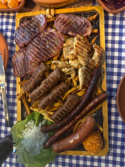 A rectangular platter of grilled meats and fries sits on a blue and white checkered surface. The platter includes grilled steak, chicken pieces, sausages, and cevapi (skinless sausages). Fries are spread throughout the platter. There is a scoop of chopped onion on lettuce leaves and a breaded ball on the side. Other food dishes are partially visible at the platter's edges.