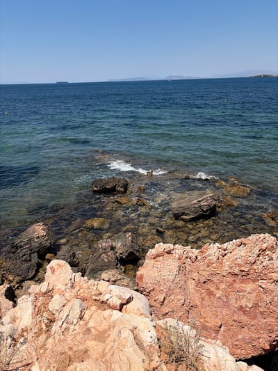 The image captures a scenic view of the sea and rocky shoreline in Vouliagmeni, Greece. The scene is divided horizontally, with the calm, blue sea on the left and the rugged, reddish-brown rocks on the right. The water is clear, allowing a glimpse of the submerged rocks beneath its surface, and gentle waves are seen lapping against the shore.
In the distance, the faint outline of mountains can be seen against the clear blue sky, suggesting a sunny day, likely in the late afternoon or early morning due to the warm light illuminating the rocks. A small boat or buoy is visible in the water on the left side of the frame, and a larger ship can be seen on the horizon. A patch of dry, sparse vegetation grows on the rocks, adding to the arid, Mediterranean feel of the location.
The overall mood is serene and natural, highlighting the beauty of the coastal landscape. There are no people or animals visible in the frame. The textures of the water and the rocks are a prominent feature, with the rough, layered appearance of the rock contrasting with the smooth, rippling surface of the sea.