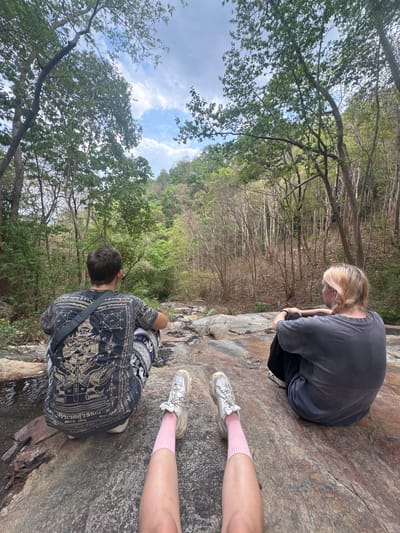 Two people are seated on a rock overlooking a wooded area in Suthep, Thailand. A person with blond hair wearing a gray t-shirt sits with their head bowed and arms around their legs. Another person sits facing away from the camera, wearing a patterned black t-shirt and carrying a gray shoulder bag. Another set of legs wearing pink socks and sneakers are also on the rock. The trees are lush with green foliage, and patches of blue sky are visible through the branches.