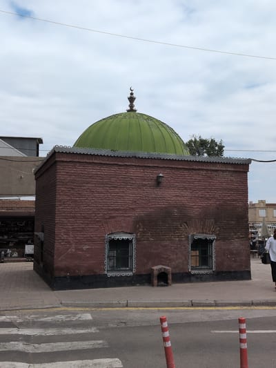 The image captures a red brick building with a green dome, located in the city of Ganja, Azerbaijan. The building has two windows with decorative frames and a small, arched niche near the base of the wall. A crescent moon finial adorns the apex of the dome. The building is situated on a sidewalk next to a street with a pedestrian crossing. In the background, other buildings and a person walking away from the camera are visible. The sky is overcast with clouds.