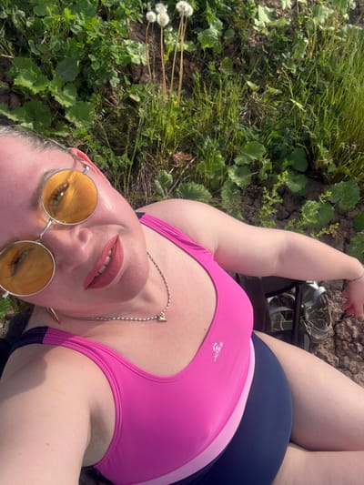 A slightly high-angle, outdoor shot shows a person lying on the ground, likely in a garden or grassy area. The person is wearing a fuchsia-colored tank top with the "JOSS" logo visible on the chest, and dark blue shorts or a swimsuit bottom. They also have on yellow-tinted, round sunglasses and a silver chain necklace with a small spade-shaped pendant. Their lips are painted with a dark pink or red lipstick, and their mouth is slightly open, showing teeth. One hand is visible near the top right, and a silver bracelet is on the wrist.

The background is a mix of green grass, various plants, and exposed soil. There are some white, fluffy dandelion seeds visible on the left side. The lighting suggests it is daytime, with bright sunlight creating highlights on the person's skin and casting some shadows. The person's expression appears relaxed or content. A pair of sandals or shoes can be seen in the upper right background, resting on the ground near some dried earth. The overall setting evokes a casual, warm-weather outdoor scene in Sapunov Bor, Russia.