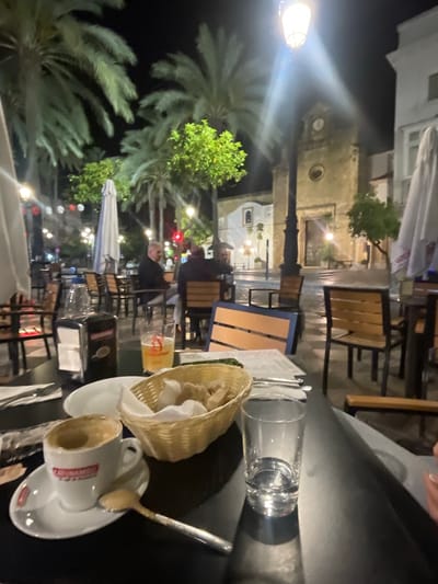 The image depicts an outdoor dining scene in Jerez De La Frontera, Spain, at night. The view is from a table, with a half-empty coffee cup on a saucer, a bread basket, a spoon, and a glass of water in the foreground. Tables and chairs are visible in the middle ground, occupied by people dining. In the background are palm trees, streetlights, and architectural structures, including a building with a clock. Umbrellas are present, suggesting an outdoor seating area. The scene is illuminated by artificial lights.