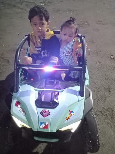 Two children are in a light blue and black toy off-road vehicle. The older child, a boy with short, dark hair, is wearing a yellow and navy blue Adidas-branded shirt. The younger child, likely a girl, has her hair in a topknot and is wearing a blue and white striped shirt. The toy vehicle has several stickers, including "Transworld Motocross" and "NCO," as well as functional headlights. The location appears to be an outdoor setting, potentially in a city area of Nganjuk, Indonesia, though specific landmarks are not visible. The surface under the vehicle is a gray, possibly paved area, with some scattered debris.
