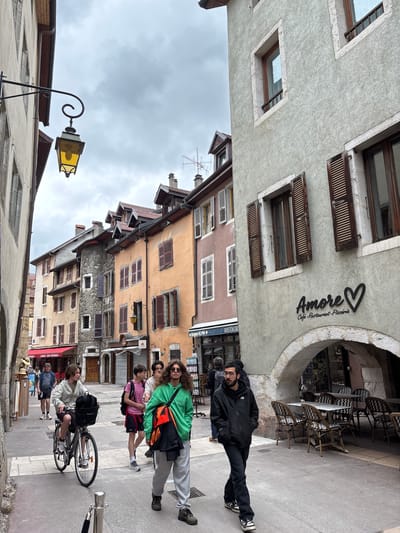 In Annecy, France, a narrow street is lined with buildings of various heights and colors. The street is paved, and there are tables and chairs set up outside a cafe on the right side of the image. The sky is overcast.

Several people are walking down the street. On the right, a man in a black jacket and dark pants walks towards the viewer, his hands in his pockets. Behind him, a group of people are walking in the same direction. One person, wearing a green hoodie, is talking to another person. To the left, a young person rides a bicycle with a basket on the front. Further down the street, more people are visible, as are shops with awnings. A street lamp with a yellow glow hangs from a bracket on the left side of the street. The architecture is typical of old European towns, with multi-story buildings featuring shutters and ornate details.
