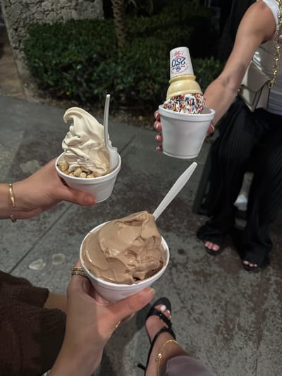The image captures a group of three people standing outside with ice cream. One person holds an ice cream cone with sprinkles, another holds a cup of vanilla ice cream with nuts, and the third holds a cup of chocolate ice cream. The location appears to be outdoors, possibly a sidewalk or patio. The image was taken in Boca Raton, United States.