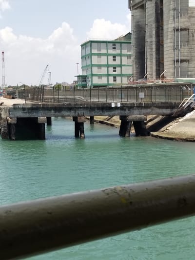 The image displays an industrial waterfront scene in Mkomani ward, Kenya, under a bright, partly cloudy sky. A blurred, dark grey metallic railing occupies the immediate foreground, running diagonally across the bottom.
In the midground, a substantial concrete pier or walkway extends over calm, turquoise-green water. This structure is supported by multiple thick, weathered concrete pillars partially submerged. The surface of the pier features a dark metal fence running along its edge, behind which a flat, paved area is visible. To the right, an angled concrete embankment slopes down into the water, with a section of white and blue stairs visible ascending from it.
The background reveals an industrial complex. A multi-story building with distinctive light green horizontal banding on white sections, interspersed with windows, stands directly behind the pier. To its right are several tall, grey, cylindrical concrete silos, characteristic of a bulk cargo facility. Further in the distance, multiple large industrial cranes, including one prominent red crane on the left and other lighter-colored cranes, are visible, indicating an active port environment. No individuals are present in the frame.