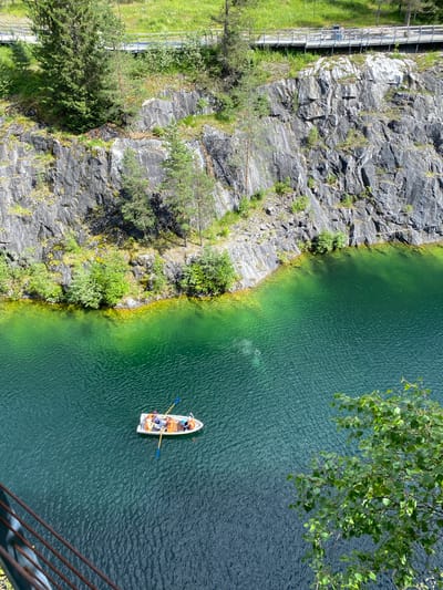 The image captures a scenic view of the Ruskeala Marble Canyon in Ruskeala, Russia. A small white boat with an orange awning is on the vibrant green water, with several people visible inside. Two individuals are actively rowing with blue oars. The boat is positioned in the lower center of the frame. Surrounding the water is a steep, rocky cliff face, characterized by grey stone with patches of green vegetation and moss. The cliff is dotted with trees, some of which are tall and slender, while others are smaller and bushier. In the upper part of the image, a wooden walkway with railings is visible, situated atop the cliff, with more greenery beyond. The water itself appears calm with a rippling texture. A leafy tree branch extends into the frame from the lower right corner, partially obscuring the view of the water. The overall lighting suggests a sunny day.