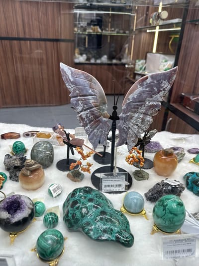 This image captures a retail display of various mineral and crystal items, likely in a shop in Bolshoye Savino, Russia. The central and most prominent subject is a large, intricately carved butterfly made from a patterned stone, possibly jasper or agate, with wings displaying shades of grey, purple, and brown with white inclusions. It is mounted on a black metal stand.
Around the butterfly, a diverse collection of polished and raw minerals is arranged on a soft, white display surface. Notable items include multiple pieces of green malachite, presented as spheres (some in golden stands) and a larger, irregularly shaped sculpture. There is also a light blue/grey sphere in a golden stand, an amethyst geode displaying purple crystals, and various other tumbled stones in hues of orange, brown, and clear. Small decorative clusters resembling orange berries on black stems are interspersed among the minerals.
Reflections on the left side of the image suggest the display is enclosed within a glass case, showing blurred views of wooden paneling and interior lighting fixtures of the shop.
Visible text on the price tags and labels provides specific information. One label identifies the vendor as "ИП Феофилов Д.Г." and the item as "Бабочка на мет. подставке" (Butterfly on a metal stand), with a price of "21 380,00 руб. за шт" (21,380.00 rubles per piece). Another price, "16 618.00 руб", and the country "РООССИЯ" (RUSSIA) with a date "28.11.2025" are also legible. The image depicts a static scene of goods for sale in an indoor retail setting.