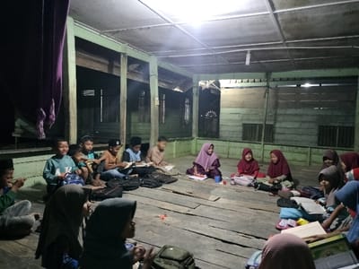 A group of approximately 15 children are seated on a wooden plank floor within a simple, semi-enclosed wooden structure at night in Cutneuheun, Indonesia. The structure features light-green painted wooden plank walls and exposed ceiling joists from which a single white fluorescent tube light hangs, providing primary illumination. Approximately six to seven boys, dressed in t-shirts and pants, are visible on the left and center, some examining books while others look towards the front or at their peers. Approximately eight to nine girls, wearing various colored hijabs and long-sleeved clothing, are primarily located on the right and in the foreground, seated around open books and occasionally interacting or smiling. Several school bags, books, and a pair of orange-handled scissors are scattered on the floor among the children. A dark purple fabric is visible hanging on the far left. The scene depicts a communal study or learning session under artificial light.
