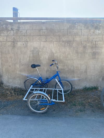 A blue tricycle with a white frame is parked against a cinder block wall in an outdoor setting. The tricycle has a seat, handlebars, pedals, and wheels, with a cargo area constructed from white tubing at the rear. The wall is made of large, rectangular cinder blocks and shows signs of wear. The ground in front of the wall is a mix of gravel and dry grass. The sky above is clear and bright. The scene appears to be located in a rural or semi-urban area.