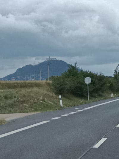 The image shows a landscape with a road in the foreground, a field and trees in the mid-ground, and a hill with a castle on top in the background. The sky is overcast. The road has white lane markings. There are some white markers along the side of the road, and a round white sign on a pole. The hill in the background is covered in trees and vegetation, and the castle appears to be made of stone. The location is Bělá pod Bezdězem, Czechia.