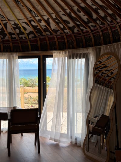 A vertical, medium shot depicts the interior of a yurt located in Chok Tal, Kyrgyzstan. The ceiling features a traditional circular structure made of wooden poles radiating outward, covered with patterned fabric. A large window with sheer white curtains reveals a view of a blue lake and grassy hills. A wooden chair sits near the window, and a decorative mirror hangs on the wall, reflecting the yurt's ceiling. The floor is made of light-colored wood.
