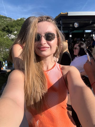 A young woman with long blonde hair, wearing sunglasses and an orange and white patterned top, takes a selfie outdoors. She is smiling and has glossy lips. Behind her, a blurred background shows a mountainous landscape with green trees under a clear blue sky, a shaded outdoor seating area with people, and a table with bottles of water. The woman's arm is visible in the foreground, resting on her shoulder. The scene suggests a casual social gathering in a scenic location.