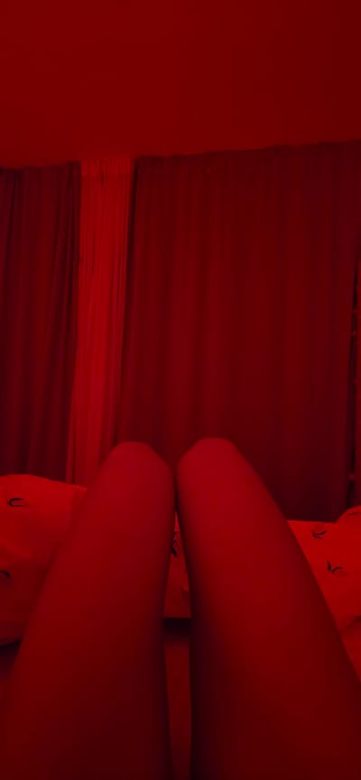 The image depicts a close-up view of a person's legs, illuminated by red light. The legs are positioned in the foreground, with the thighs appearing smooth and shapely. The lighting casts a strong red hue over the entire scene, obscuring finer details and creating a dramatic atmosphere. Behind the legs, a blurred background suggests a domestic setting, possibly a bedroom, with hints of red curtains and what appear to be pillows with a subtle pattern. The overall impression is one of intimacy and mystery, with the red light contributing to a mood that could be interpreted as sensual or even unsettling. There is no discernible indication of the city of Lieli, Switzerland, or any other specific urban location. The focus is entirely on the subject's legs and the surrounding red-lit environment.