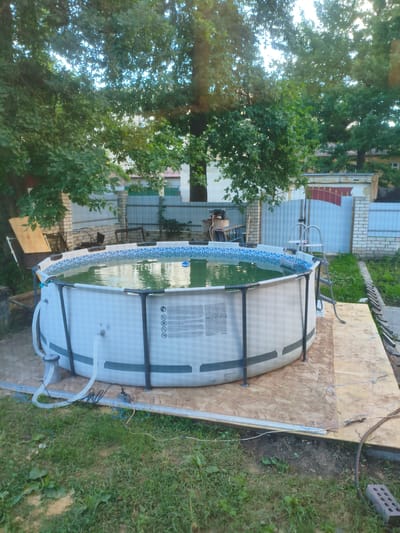 This is a medium shot, outdoor, vertical-oriented photograph taken on a sunny day, likely in the afternoon given the light. The scene is a backyard in Kotovsk, Russia, featuring a large, round, above-ground swimming pool filled with murky green water. The pool has a white exterior with a blue and white patterned rim. A silver ladder is attached to one side of the pool. The pool is situated on a flat platform made of large sheets of light-colored plywood.
Around the pool, there is lush green grass. In the background, behind a light-colored brick fence, a blue metal gate is visible, and beyond that, residential buildings and more trees. The overall setting suggests a suburban or semi-rural environment.
There are no people visible in the image, but the presence of the pool, ladder, and electrical connections suggests it is set up for use. The water condition implies it may be in need of maintenance or has been recently filled. The time of day is suggested by the bright sunlight casting shadows, typical of a clear afternoon. The weather appears to be warm and dry.
Visible text on the pool's exterior includes warning symbols and what appears to be the brand name "BESTWAY CORP.COM". The scene conveys a sense of summer leisure, though the pool's condition adds a slight element of disarray or a work-in-progress feel.