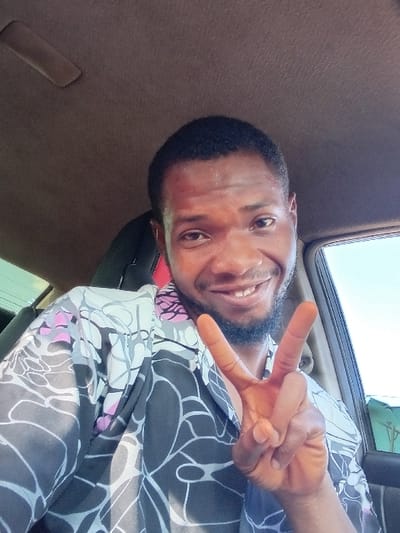 A man with dark hair and a beard smiles, holding up a peace sign in the passenger seat of a car. He is wearing a patterned shirt with dark, grey, and white colors, along with touches of pink. The interior of the car is brown. A window is visible, showing a bright blue sky and the top of a green structure.