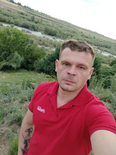 A man in a red polo shirt with a Coca-Cola logo is taking a selfie outdoors. He has short, light brown hair and light blue eyes. He is looking directly at the camera with a neutral expression. A tattoo of a bird is visible on his left arm. The background is a lush green landscape with trees and a river, suggesting a rural setting. The sky is overcast, indicating it might be early morning or late afternoon. The location is identified as Koksayaq rural district in Kazakhstan. There is no specific activity or event taking place, it appears to be a casual portrait.