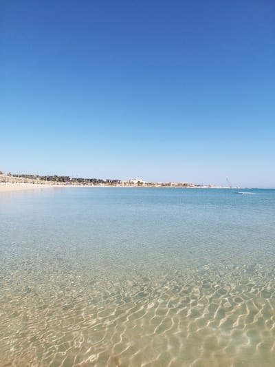 This tranquil wide shot captures a serene coastal scene in Safaga, Egypt, under a vast, cloudless blue sky. The foreground is dominated by incredibly clear, shallow turquoise waters, allowing a visible view of the sandy seabed below, marked by shimmering light patterns from the gentle ripples on the surface. The water gradually deepens further out, maintaining its calm, reflective quality.

To the left, a long, light-colored sandy beach stretches along the shore, appearing mostly empty with just a few straw umbrellas and sun loungers visible, suggesting a peaceful, uncrowded atmosphere. Along the shoreline, in the mid-ground, a series of modern, light-colored buildings, characteristic of a resort or hotel complex, are nestled among scattered palm trees.

In the far distance to the right, a large industrial crane can be seen near the water, possibly indicating port activity or ongoing construction. A flat, white floating platform is also visible further out in the calm sea, along with a small, indistinct dark object.

The bright, even illumination suggests a sunny daytime, likely mid-morning or early afternoon, with perfect clear weather. The overall impression is one of peaceful relaxation and natural beauty, characteristic of a vacation destination. No people or visible text are discernible in the image.