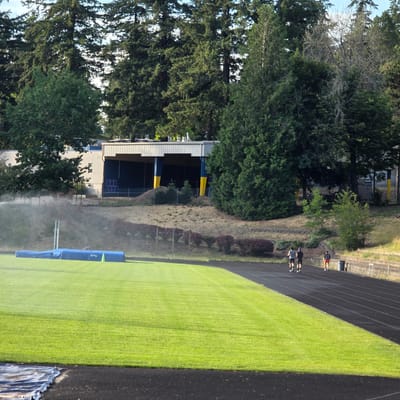 An outdoor track and field scene is depicted, likely in Portland, United States, given the lush greenery and architecture consistent with the region. In the foreground, a vibrant green lawn meets a black running track. To the left, a blue landing pad for pole vaulting is visible, with a pole vault standards set up. A sprinkler system is actively watering the grass, creating a visible mist.

In the midground, three individuals are running on the track. Two are running side-by-side in the direction away from the camera, wearing dark athletic wear. A third individual is slightly ahead, also running away from the camera, wearing red shorts and a dark top. A trash can is positioned next to the track near this individual.

The background features a building with a gray, corrugated metal roof and a blue and yellow facade, possibly a stadium or gymnasium. Behind the building and track, a dense forest of tall evergreen trees dominates the landscape, with some deciduous trees interspersed. The ground on the hillside leading up to the building is covered in dry grass and scattered bushes. The sky above is bright and clear, indicating daytime.