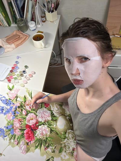 A person wearing a facial mask and a grey tank top is painting a floral picture at a desk in an indoor setting. The desk has various art supplies, including small paint containers, a brush, and a cup of tea. The background shows shelves filled with books and art materials. The person is focused on the painting, holding a brush in their right hand. The lighting is artificial, suggesting an indoor environment. The image is taken from a slightly high angle, looking down at the person and their work. The person's face is partially obscured by the facial mask, but their eyes are visible and looking towards the camera. The overall scene depicts a moment of creative activity in a personal space.