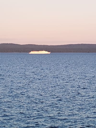 The image shows a large white ferry boat sailing on a calm, blue body of water. In the background, a dark, tree-covered hill or landmass is visible under a soft pink and orange sky, suggesting either sunrise or sunset. The water's surface is rippled, creating a textured effect. The ferry is positioned horizontally across the frame, its long form illuminated by the sky's light. No people are visible in the image. The scene appears to be outdoors in a natural setting, possibly a lake or a calm sea. The location is identified as Kakkarovo, Russia.