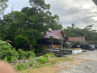 The image shows a small restaurant called "KUAH CUE & Manok Budee" located in the city of Seungkomulat, Indonesia. The restaurant appears to be a wooden structure with a reddish-brown roof, set amidst lush green trees and foliage. A sign above the entrance advertises the restaurant's specialties, "Kaki Kulu" and "Khas Aceh Raya". In front of the restaurant, there is a parking area with several cars visible, including a black SUV. The sky is overcast, suggesting a cloudy day. The area around the restaurant is natural and somewhat unkempt, with grass, rocks, and branches scattered on the ground. The overall impression is of a rustic, local eatery situated in a natural environment.