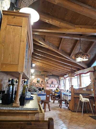 The image depicts the interior of a rustic restaurant, likely in Pontagna, Italy, with a focus on its wooden architecture and ambiance. A large, dark wooden cabinet dominates the left side of the foreground, its doors partially open, revealing glass panes. Several bottles, appearing to be olive oil or wine, are neatly arranged on a speckled countertop in front of the cabinet.

In the background, diners are seated at tables covered with blue and white tablecloths. A man wearing a yellow and blue striped shirt sits at a table, facing away from the camera. Another person is seated opposite him. The restaurant features a sloped wooden ceiling with exposed beams, and decorative hanging lamps with intricate metalwork. Large windows with red and white patterned curtains allow natural light to enter the space. The floor is tiled with terracotta-colored squares. A white high chair is positioned near one of the wooden booths. Shelves on the back wall display bottles and decorative items. The overall impression is that of a warm, traditional dining establishment.