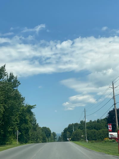 The image shows a straight asphalt road passing through a rural area with trees lining both sides. The sky is a clear blue with scattered white clouds. Power lines run along the right side of the road, supported by wooden poles. A Canadian flag is attached to one of the poles, near a red mailbox. The area appears to be residential, with manicured lawns and gardens visible. The overall scene depicts a serene summer day in a Canadian countryside setting.