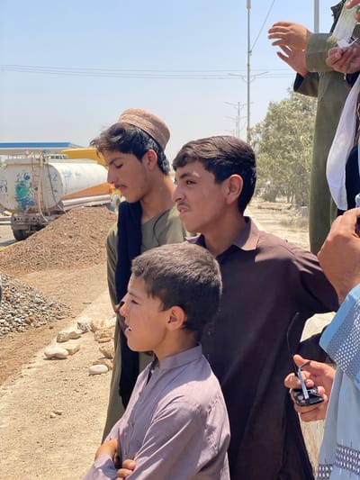 In the city of Kakar, Afghanistan, three young boys are pictured standing in an outdoor environment. The boy on the far left, wearing a brown embroidered cap and a dark green shirt with a black scarf, is looking down and to the left. The boy in the center, wearing a brown shirt, is looking to the right with his mouth slightly open. The boy on the right, wearing a light purple shirt, is also looking to the right. In the background, a water truck is visible with a pile of gravel and rocks. The ground is covered in dirt and small stones. The sky is clear and blue.