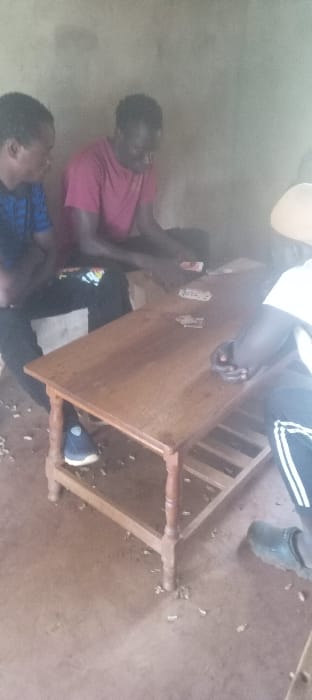 Three men are observed indoors at a location identified as Getenga ward, Kenya. They are seated around a rectangular wooden table with turned legs and a lower shelf structure. The table is centrally positioned on a light brown, possibly earthen or concrete, floor, which has scattered light-colored debris. The background consists of a plain, light brown wall.
The individual on the left wears a blue patterned short-sleeved shirt and dark pants, with a dark blue shoe visible on their left foot. They are looking towards the table. The person in the middle wears a red short-sleeved t-shirt and dark pants; their hands are near the table surface. The individual on the right is partially visible, wearing a white shirt and dark pants with white stripes, and a dark shoe or clog. This person's hands are holding a fan of playing cards.
Multiple playing cards are spread across the wooden table surface, indicating a card game in progress. All three individuals are focused on the table or the cards.