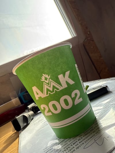 The scene is an interior view, likely a desk or table surface, located in Hrishteni, Bulgaria. The primary subject is a green paper cup, prominently positioned in the center. The cup features a white logo with a stylized 'M' topped by a crown or flame, above which are the Cyrillic letters "АМК" and the number "2002". Two white horizontal stripes encircle the lower portion of the cup, and its rim is white.
The cup rests on light-colored documents, which display visible Cyrillic text, including words such as "БАНКОВАТА", "ОКОЛНАТА", "МИНИСТЕРСТВО НА", and "УСЛОВИЯ". To the right of the cup, a black object resembling a car key fob or remote control with silver accents and a small green detail is visible. To the left of the cup, a dark, angular object with a cylindrical component, possibly part of an electronic device or tool, is present. Further left, indistinct objects include what appears to be a blue container and a reddish item.
Bright light originates from a window or light source positioned in the upper left quadrant of the image, creating highlights and shadows within the scene. The upper right background consists of a dark, textured surface, likely a wall or furniture. No individuals are depicted, and no specific actions or interactions are occurring. The environment suggests an indoor setting, possibly an office or personal workspace.