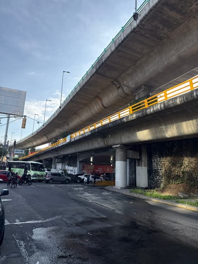 A wide shot captures a sunlit day in Tizapan, Mexico, beneath a prominent elevated highway. The concrete structure, painted with a green railing on top and a bright yellow one below, dominates the upper half of the frame, casting shadows on the street and lower level. The street level is busy with traffic, including a green bus, several cars, and a motorcycle with riders. People are seen walking and crossing the street. The road surface is wet and reflects the light. Greenery grows along the embankment next to the highway's support columns. A billboard is visible on the left side of the image, and banners with text are displayed on the lower level of the highway structure.