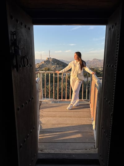 A female subject is observed standing on a wooden balcony, framed by an open, weathered wooden doorway, overlooking the mountainous landscape of Hatta, United Arab Emirates. The doorway, positioned in the foreground, features visible dark metal studs and a padlock attached to a metal ring on its left panel.
The subject, with long dark hair, is attired in a white long-sleeved hooded sweatshirt, matching white sweatpants, and white sneakers, carrying a light-colored crossbody bag. She stands with her arms extended, hands resting on a white-painted metal railing that encloses the balcony. Her posture is relaxed, and her head is turned towards the left, appearing to observe the expansive view.
The environment is illuminated by late afternoon sunlight, casting long shadows across the wooden plank flooring of the balcony. Beyond the railing, the middle ground reveals a prominent rocky hill topped with two communication towers and a stone structure. Below this, a dense grove of date palm trees is visible. The background consists of rugged, arid mountains under a clear blue sky that transitions to a warmer hue near the horizon. Distant signs of urban development are faintly discernible amidst the mountains.